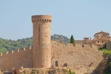 Tahkim edilmiş bir duvarla çevrili, uzun, yuvarlak bir kulesi olan tarihi bir taş kale. Kale, yeşil tepelerden oluşan bir zemin ve bazı ziyaretçilerin görebileceği açık mavi bir gökyüzüne karşı kurulmuş..