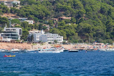 Suda tekneleri olan, yemyeşil tepeler ve konut binalarıyla çevrili manzaralı bir sahil manzarası. Kumsal güneş banyosu ve şemsiyelerle dolu..