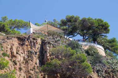 Uçurumun manzarası, yemyeşil ve modern bir yapı. Bina geniş bir gölgeliğe sahiptir ve açık mavi gökyüzüne bakan ağaçlarla çevrilidir..