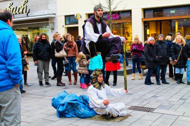 Almanya, Düsseldorf - 21 Aralık 2013 - Her ikisi de özenle hazırlanmış kostümler giymiş bir başka sanatçı aşağıda otururken yerden yükselen bir sokak sanatçısı. Kalabalık şaşkınlıkla izliyor, arka planda dükkanlar var..