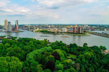 Modern binalar ve kıyı boyunca uzanan yemyeşil parklar arasında kıvrılan bir nehrin hava manzarası. Tekneler suda görülür ve ufuk çizgisi parçalı bulutlu bir gökyüzünün altında mimari bir karışım içerir..