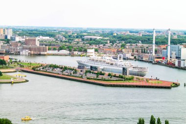 Yanaşmış büyük bir yolcu gemisinin liman manzarası. Çevresindeki bölgede modern binalar, yeşil alanlar ve bir nehir bulunur. Arka planda, endüstriyel yapılar ve bir ufuk çizgisi bulutlu bir gökyüzünün altında görülebilir..