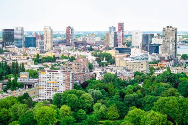Yüksek binalar ve yeşil parkların yer aldığı modern şehir siluetinin panoramik bir görüntüsü. Kentsel manzara konut ve ticari yapıların bir karışımını sergiliyor, ağaçlar verimli bir ön plan sağlıyor.