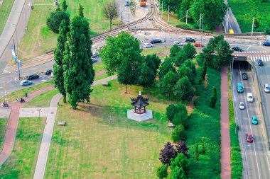 Çimenlerle ve yemyeşil ağaçlarla çevrili geleneksel bir taraçanın bulunduğu bir parkın havadan görünüşü. Yakınlardaki yollar, arabalı ve kavşaklı trafiği gösteriyor. Sahne, doğa ve şehir altyapısının bir karışımını yakalıyor..