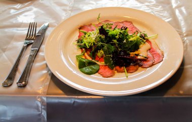 Dilimlenmiş biftek, taze yeşillikler ve beyaz bir tabakta zarif bir şekilde hazırlanmış kremalı bir sos içeren bir gurme tabağı. Çatal bıçak takımı tabağın yanına plastik kapaklı bir masanın üzerine konur..