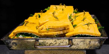 Yeşil otlar ve kürdanlarla süslenmiş, düzgünce hazırlanmış sandviçlerle dolu büyük bir tepsi. Sandviçler kaymak gibi görünüyor ve kırsal bir yüzeye yerleştiriliyor..