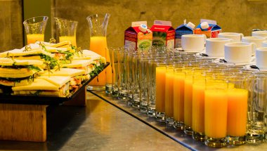 İçinde bir tabak sandviç, bir bardak portakal suyu ve bir kutu süt bulunan bir büfe. Sandviçler yeşilliklerle düzgünce düzenlenirken, portakal suyu berrak bardaklarda servis ediliyor, canlı ve davetkar bir vitrin oluşturuyor..