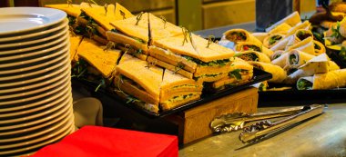 Düzenli sandviç ve paketlerin yer aldığı bir açık büfe. Sandviçler görünür yeşilliklerle dolu ve kürdanlarla bir arada tutuluyorlar. Yakınlarda, sarılmış sargılar var, yayılıma çeşitlilik katıyor. Tabaklar ve kırmızı peçeteler de mevcut., 