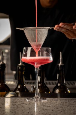 Bir barmen bardağa kokteyl dökerken ince bir ağ süzgeci kullanır. Bar ortamında profesyonel miksoloji teknikleri sergiler..