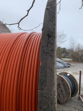 Kırmızı-turuncu renkli elektrik kablosu olan büyük bir makaranın ucuna yakın çekim. Bir inşaat alanının arka planında siyah kablo ile küçük bir ahşap makara var. Yan görünüm.