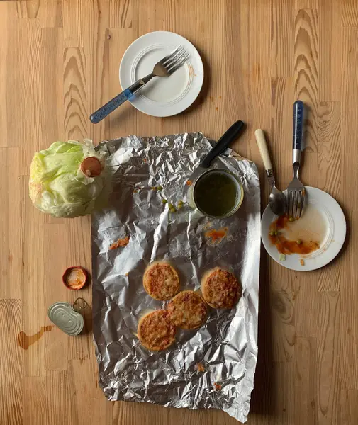 Yemekten sonra eski mutfak ahşap masasında kirli bulaşıklar var. Folyo, buzdağı lahanası, iki kirli beyaz tabak, çatal, bıçak, çay kaşığı, teneke kutu, domates sosu, mini pizza, kapak. İki kişilik natürmort akşam yemeği..