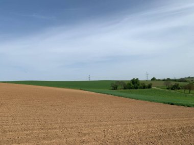 Açık mavi gökyüzünün altında yeni sürülmüş bir tarlanın yer aldığı manzaralı bir kırsal alan. Ön plandaki kahverengi toprak, arka plandaki yeşil alanla çelişiyor ve derinlik ve açıklık hissi yaratıyor. Elektrik hatları ve direkler Dist 'te görülebilir.