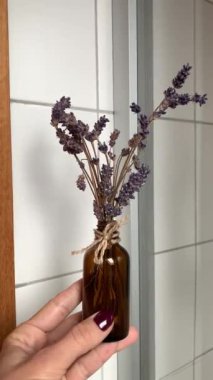 Doğal sicim yayı ile bağlanmış kurutulmuş lavanta saplarıyla dolu kehribar rengi küçük bir cam şişenin rahatlatıcı bir sahnesi. Arka planda, rahat ve spa benzeri bir atmosfer ekleniyor..