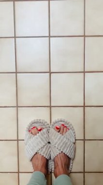 Ayaklar rahat, terlikler bej kaplamalı zeminde. Kırmızı boyalı ayak tırnakları renk katar. Minimalist ev konforu ve rahatlama konsepti