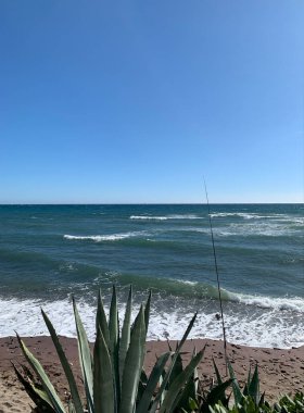 Endülüs, İspanya 'nın Costa del Sol plajından çarpıcı okyanus manzarası. Sahilde bir olta kamışı duruyor, ön planda büyük sularla, sakin bir kıyı manzarası yaratıyor.