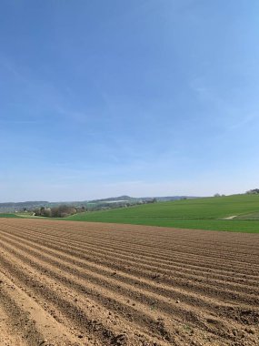 Ön planda yeni sürülmüş tarlaları ve açık mavi gökyüzünün altında yemyeşil tepeleri olan manzaralı kırsal bölge. Orta Avrupa 'da tipik bir bahar mevsimi için huzurlu bir kırsal manzara.