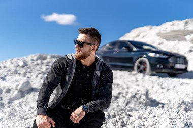 Açık havada arabayla poz veren maço bir model. Güneş gözlüklü yakışıklı genç adam..