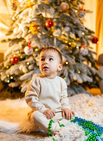 Cute child with christmas tree. Happy baby sitting near a fir tree and holding a christmas ornament and smiling. Merry Christmas and Happy Holidays!