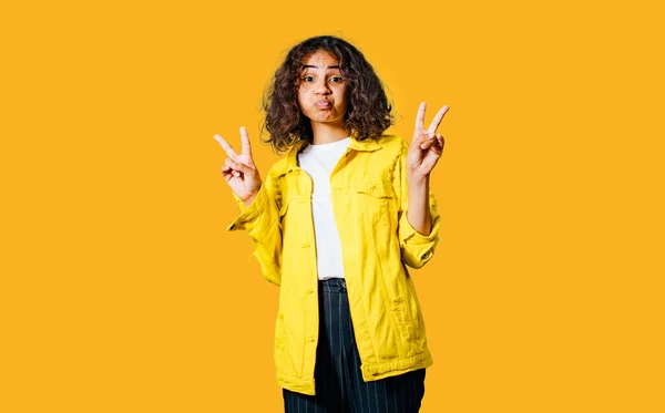 Positive human emotions. Headshot of happy emotional teenage girl with bob curly haircut with hands making peace sign, wearing bright makeup. Blue background. Wearing bright yellow cotton jachet and white shirt. Funny emotion of duck face. Yellow bac