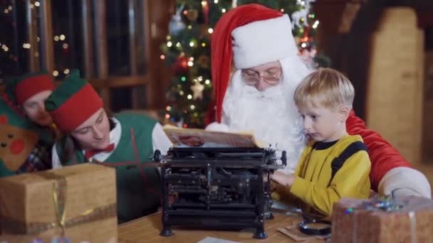 少年はサンタに願い事を書いて手紙を書く サンタクロースとエルフの近くの古いマシン上の小さな男の子の印刷文字 — ストック動画