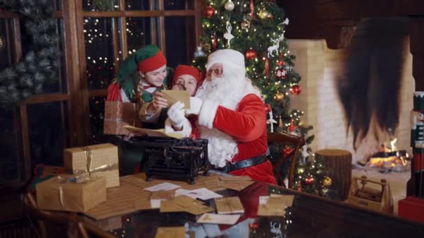 圣诞老人在看孩子们的邮件圣诞老人和精灵坐在桌旁 在房间里看书 — 图库视频影像