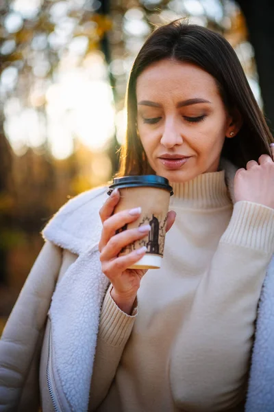 Young woman smelling coffee in autumn park. Outdoor female person enjoying coffee.