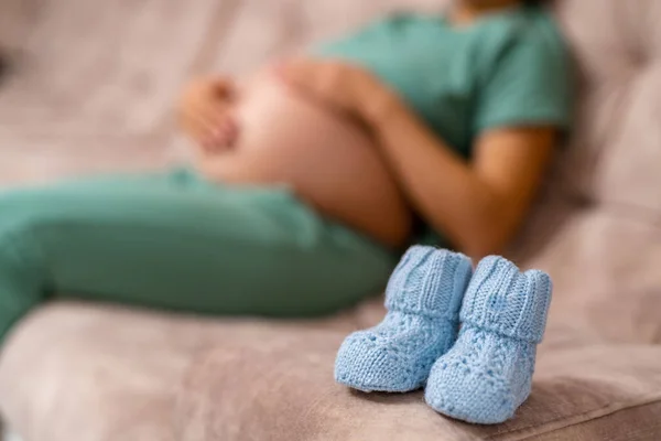 A baby resting next to a pair of blue shoes on a cozy couch. A baby laying on a couch next to a pair of blue shoes