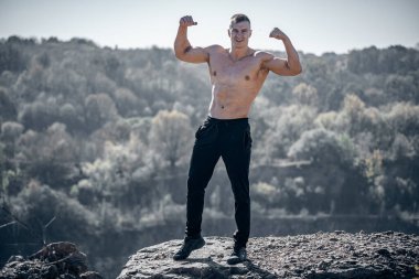 Bir kayanın üzerinde üstsüz bir adam duruyor. Rocky Cliff 'te Zaferle Duran Adam