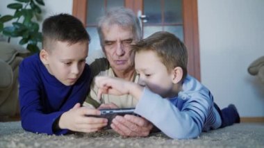 Çocuklu yaşlı bir adam video oyunu oynuyor. Ergen çocuklar dedenle evde yerde yatarken akıllı telefondan yazı yazıyorlar.