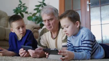Aile teknolojisini evde kullanıyor. Mutlu yaşlı adam ve torunları cep telefonlarını oynarken evde yerde dinleniyorlar.