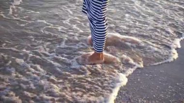 Plajda suyla oynayan bir kadın. Sudaki dişi bacaklar. Uzun elbiseli bir kadın köpüklü dalgalarda su sıçratıyor ve ayaklarıyla su oynuyor..