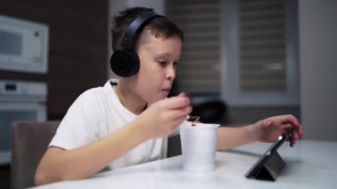 Çocuk mutfakta yemek yiyor. Evdeki masada yemek yerken siyah kulaklıklı çocuk tablet çalıyor..