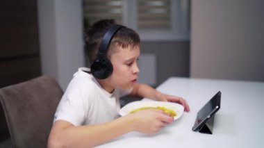 Çocuk dijital tablete bakarken evde yemek yiyor. Kulaklıklı çocuk mutfakta masada oturuyor ve yemek yiyor. Yetenekler ve çocuklar.