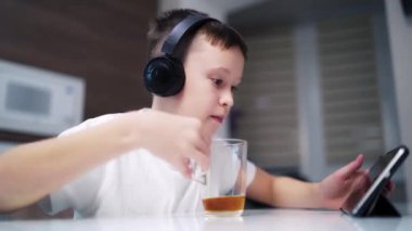 Kulaklıklı çocuk tablet ve içkiyle oynuyor. Oyuna odaklanmış genç çocuk masada oturuyor ve evde çay içiyor..