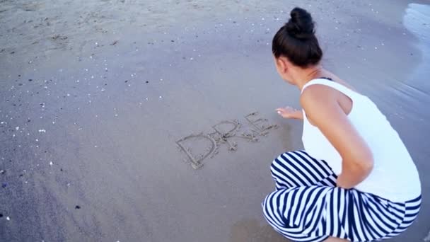 海岸の女性のバックビュー 海の近くの砂浜で夢を書く女性 水の波が書かれた言葉を洗い流しています トップビュー — ストック動画