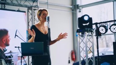 Güzel bir kadın mikrofona şarkı söylüyor. Sahnedeki düğün videosunun arka planında siyah elbiseli genç bayan şarkıcı.