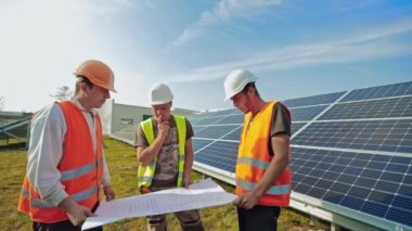 Güneş panellerinin kurulumu. Yazın bir güneş çiftliği planına bakan üç işçi. Yenilenebilir enerji alanındaki mavi güneş pilleri.