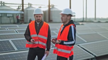 Yenilenebilir enerjinin fotovoltaik çiftliğindeki teknisyenler. Güvenlik kasklı iki işçi güneş panellerinin arka planında konuşuyorlar..