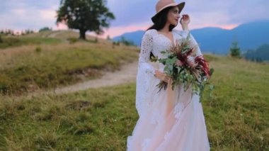 Lüks elbiseler içinde güzel bir gelin. Şapkalı ve beyaz elbiseli genç kadın elinde buket tutuyor ve gün batımında güzel doğanın tadını çıkarıyor..