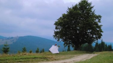 Doğada gelinlik giymiş bir kadın. Dağlardaki yeşil ağacın yanında duran beyaz elbiseli ve şapkalı genç gelin..