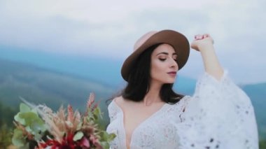 Şapkalı ve beyaz elbiseli hoş bir kadın. Koyu saçlı, elinde çiçek buketi olan ve kameraya poz veren güzel bir manken..