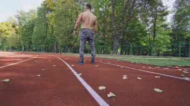 Spor koşucusu açık hava atletizm pistinde. Yeşil ağaçların arka planında stadyum yolunda koşan tişörtsüz bir sporcu. Hareket kamerası..