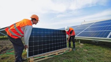 Güneş panellerinin açık havada kurulumu. Üniformalı ve kasklı işçiler güneş enerjili bir çiftlikte yeni bataryalar taşıyorlar. Yenilenebilir enerji.