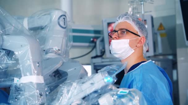 医学ロボットの近くのマスクの医者 ロボットデバイスの近くに立っている間 眼鏡と医療ユニフォームの外科医 手術コンセプト — ストック動画