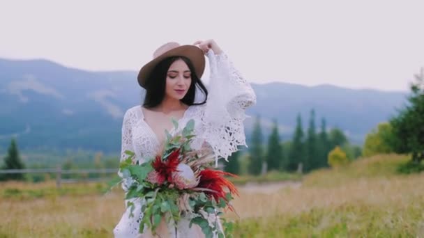 一个戴着帽子的新娘的半身画像 身着白衣的美丽而神秘的女人 在山后的背景上握着一束鲜花 — 图库视频影像