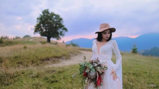 日没の自然の中で美しい花嫁 白いウェディングドレスの魅力的な女性と青い山に対して花束を保持する帽子で — ストック動画