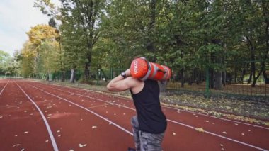Güçlü bir atlet açık havada sıkı egzersizler yapıyor. Sonbahar parkının yanındaki stadyumda omuzlarında ağırlıklarla spor antrenmanı yapıyor. Yavaş çekim.