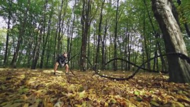 Sportif adam ormanda savaş halatlarıyla egzersiz yapıyor. Sporcu güz günlerinde antrenman yaparken kablolarla dalgalar yapıyor. Yavaş çekim.