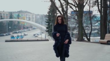 Şehir parkında kameraya poz veren çekici bir kız. Dışarıda yürüyen siyah elbiseli güzel bir manken. Kürk mantolu güzel bir kız. Yavaş çekim.