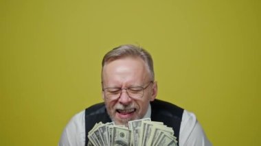 Paralı mutlu bir son sınıf öğrencisinin portresi. Gözlüklü olgun bir adam bir sürü dolar tutar ve sarı arka planda neşeyle ağzını açar. Yavaş çekim.